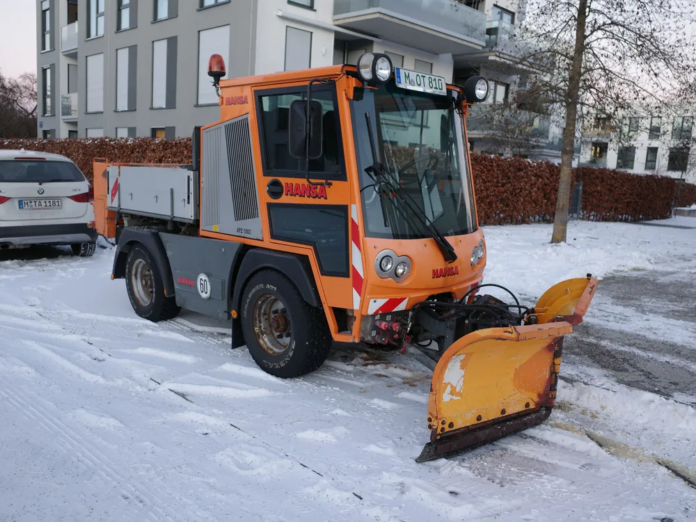 Räumfahrzeug mit Schneeschild auf einer verschneiten Straße vor einem Wohnhaus